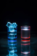 Two, blue and red shot glasses full of drinks one of wich with ice on a black background.
