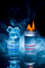 Two, blue and red shot glasses full of drinks one of wich with ice and second is burning in a thratrical smoke on a black background.
