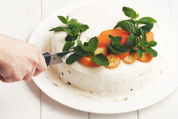 Tasty homemade cream cheese cake with fresh fruits and blueberries and mint on white plate on light wood background with knife in hand 