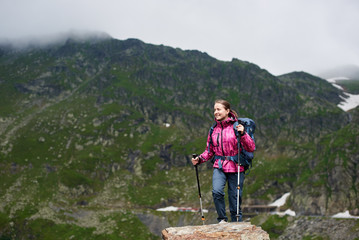 Young hiker woman climbing up in green rocky mountains with foggy clouds. Smiling girl in pink jacket and gray pants with blue backpack and trekking sticks enjoying view of wide mountain valley.