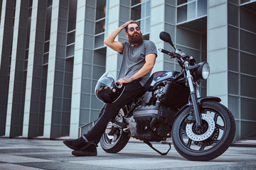 Obraz na płótnie Canvas Brutal bearded male in glasses dressed in a gray t-shirt and black pants fix the hair and holds a helmet sitting on his custom-made retro motorcycle against skyscraper.