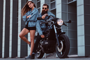 Obraz na płótnie Canvas Attractive hipster couple - bearded brutal male in sunglasses and jeans jacket sitting on a retro motorcycle and his young sensual girl standing near, posing against a skyscraper.