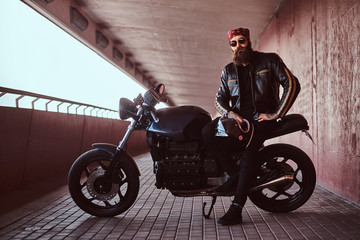 Stylish bearded male dressed in a black leather jacket and bandana with sunglasses sitting on his custom-made retro motorcycle, looking at a camera.