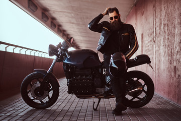 Stylish fashionable biker dressed in a black leather jacket with sunglasses fix the hair and holds...