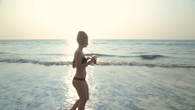 A young cute blonde woman in a swimsuit listens to music in large black headphones and dances on a tropical beach on the shore in the oncoming waves of the ocean and holds a glass of fruit juice.