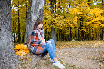 Portrait of young fashion woman outdoor. Writes in a notebook. University Park, study concept. Brunette woman in autumn park with fashionable plaid coat and scarf.