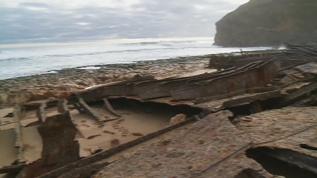 Pan right to left showing the corroded remains of a ship wreck on a sandy beach with surf in the background