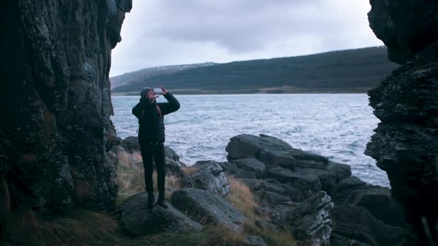 Wide shot of a young man taking photos with his cell phone while exploring rocky terrain in Iceland