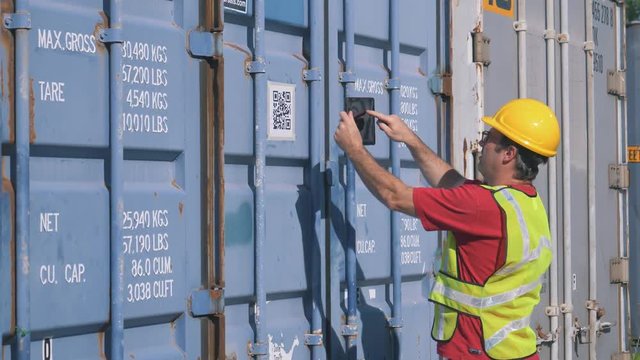 Worker using a tablet computer to scan a QR Code in a shipping yard filled with large shipping containers.