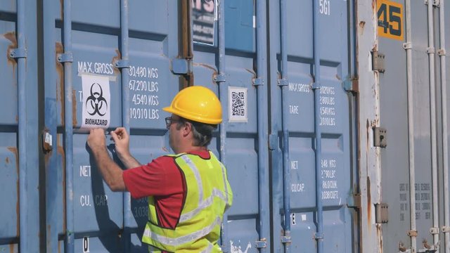 Shipping worker sticking a biohazard sign on a shipping container in a shipping yard filled with large shipping containers.