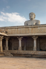 Shravanabelagola, Karnataka, India - November 1, 2013: At the Jain Tirth, gray granite Giant Bhagwan Bahubali puts torso above brown stone temple building against light blue sky.