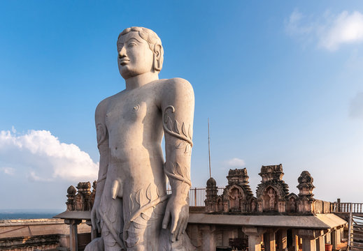 Shravanabelagola, Karnataka, India - November 1, 2013: At the Jain Tirth, gray granite Giant Bhagwan Bahubali statue at sunset. Blue sky with whie cloud. Brown stone temple building as backdrop