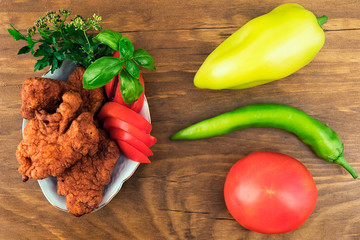Fish in the dough, deep-fried with a ruddy crust, laid on a plate with slices of tomatoes with parsley and basil , next to the whole fruit of pepper and tomato, top view.