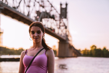 Young Italian American Woman with Long Hair and no makeup Smiling at Waterfront with Bridge and woods in background during golden hour