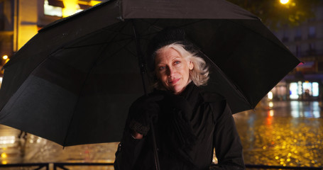 Happy senior woman in Paris smiling under umbrella on rainy night in the city