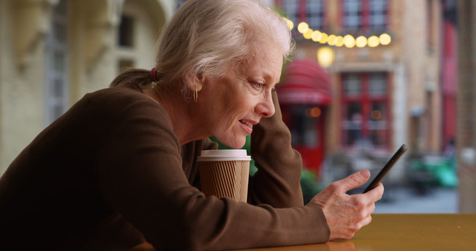 Smiling older Caucasian woman on cafe patio texting with friend on cellphone 