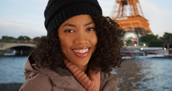 Happy black woman smiling in front of the Eiffel tower wearing jacket and beanie