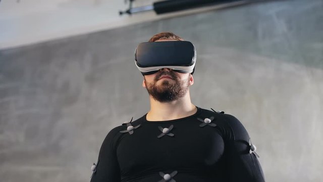 handsome bearded man in black mocap suit with motion sensors using virtual reality glasses headset on gray background futuristic interactive experience unrecognisable male player video-game simulator