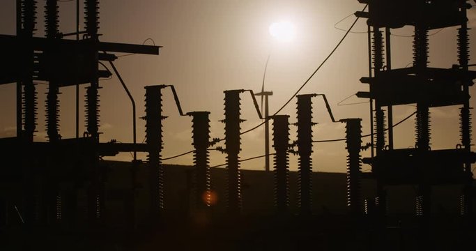 Power plant with wind tower in background silhouetted against the sun.
