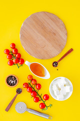 Ingredients for cooking pizza. Cherry tomatoes, olive oil, cheese mozzarella, spices near cutting board for dough on yellow background top view mockup