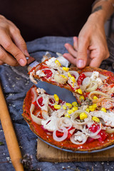 Woman hands hold a piece of raw vegan pizza with dehydrated dough, raw tomato sauce, vegan mayonnaise, corn and fresh vegetables. Healthy Italian food