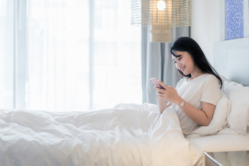 Beautiful young smiling Asian woman lying in white bed and using a phone in her bedroom..