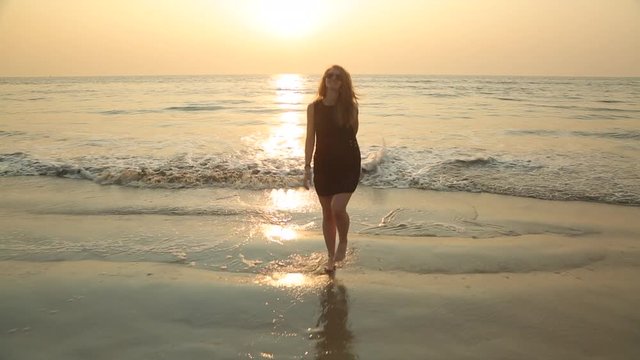 A young sexy blonde woman with long hair in a black short translucent dress and sunglasses playfully enters and leaves the sea circling in the oncoming waves and raises her hands to the sunset. 