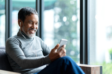 Portrait of attractive mature asian man retired with stylish short beard using smartphone sitting or listening music in urban lifestyle coffee shop. Old man using social network internet technology.