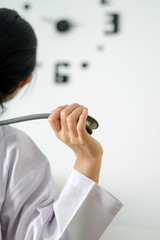 Back view of behind young female doctor holding stethoscope standing and hand up for relaxing after finish work with clock on the wall.
