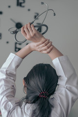 Back view of behind young female doctor holding stethoscope standing and hand up for relaxing after finish work with clock on the wall.