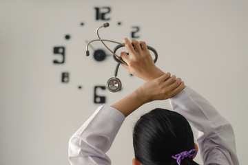 Back view of behind young female doctor holding stethoscope standing and hand up for relaxing after finish work with clock on the wall.