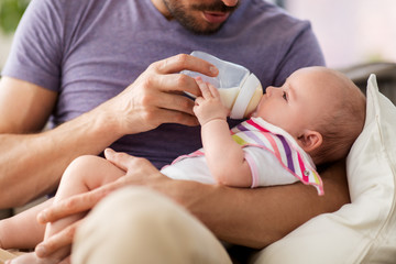 family, parenthood and people concept - close up of father feeding little daughter with baby formula from bottle at home - obrazy, fototapety, plakaty