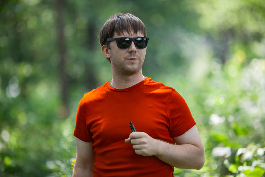 A young white bearded man in red t-shirt and in sunglasses is vaping an electronic cigarette in the forest in the summer. Close up.