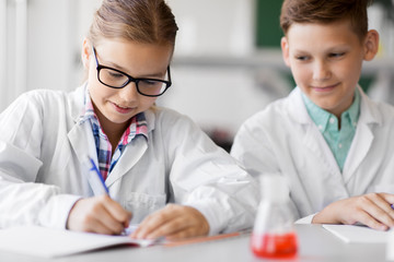 education, science and children concept - kids studying chemistry at school laboratory and writing to workbooks
