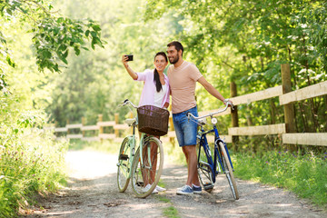 Obraz premium people, technology and lifestyle concept - happy couple with bicycles taking selfie by smartphone at summer park