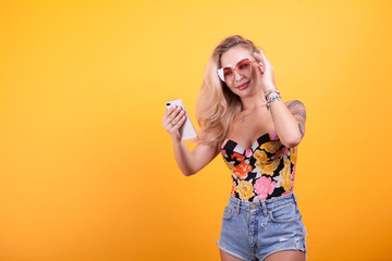 Smiling girl looking at phone screen in her hands with sunglasses in studio over yellow background