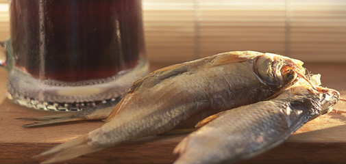 Still life with dark beer, a bottle of dark glass and three salted dried river fishes on a wooden board on the windowsill.