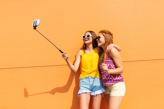 fashion, leisure and technology concept - smiling teenage girls taking picture by smartphone on selfie stick outdoors in summer