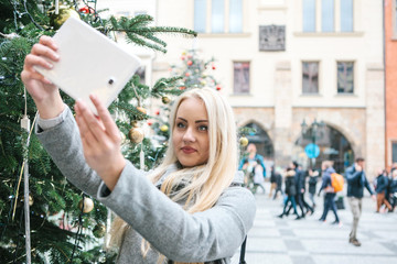 A beautiful young blonde woman or girl doing selfie or photographing next to a Christmas tree during Christmas holidays at the Old Town Square in Prague, Czech Republic.