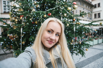 A beautiful young blonde woman or girl doing selfie next to a Christmas tree during Christmas holidays at the Old Town Square in Prague, Czech Republic.