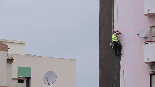 A man painting the outside of the building, hanging by cables. Painting a wall.