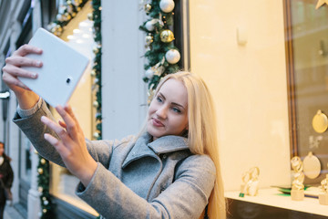 Fototapeta na wymiar A young beautiful blonde woman makes selfie next to the shop window in the time of Christmas sales.