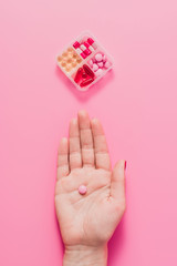 cropped shot of woman holding pink pill on pink surface with plastic container for medicines