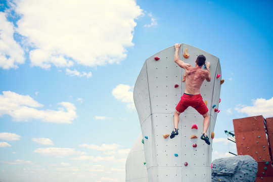 Photo from back of athlete in red shorts practicing on wall for climbing against blue sky with clouds