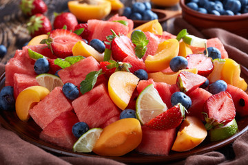 close-up of watermelon peach and berry salad with mint and lime wedges on a clay plate on a dark kitchen wooden table with ingredients, wooden spoon and napkin at the background