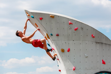 Photo of young sportsman in red shorts hanging on wall for rock climbing against blue sky with clouds