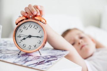 The alarm clock is on the bedside table by the bed. The little boy turns off his alarm clock. Wake up. Morning. Cozy. School.