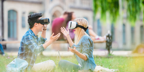 side view of multicultural students in virtual reality headsets with laptops sitting on green grass in park