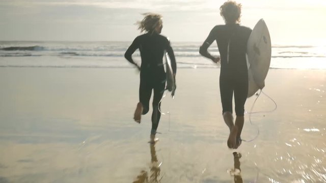 Young male surfers running from beach into the ocean waves with their surfboards to surf in the sunrise. Enjoying water sports with friends together in surfcamp on summer vacation.