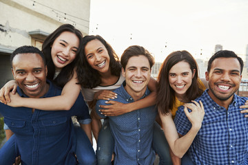 Portrait Of Men Giving Women Piggybacks On Roof Terrace With City Skyline In Background